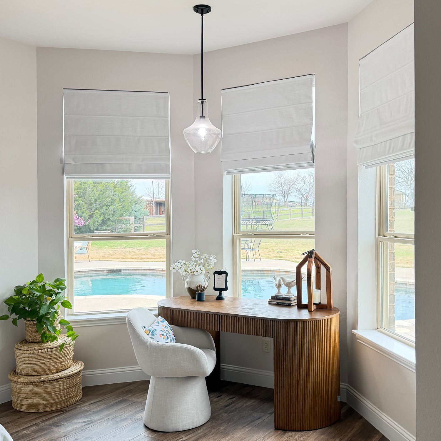 A modern home office corner featuring a curved wooden desk, a white sculptural chair, and a clear glass pendant light. The room has light gray walls, white Roman shades, and large windows looking out to a swimming pool and grassy yard.