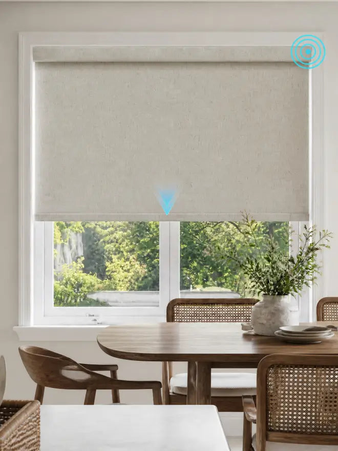 A light beige roller blind with a textured linen-like finish, partially drawn over a white-framed window in a bright, modern dining room with a wooden table and chairs.