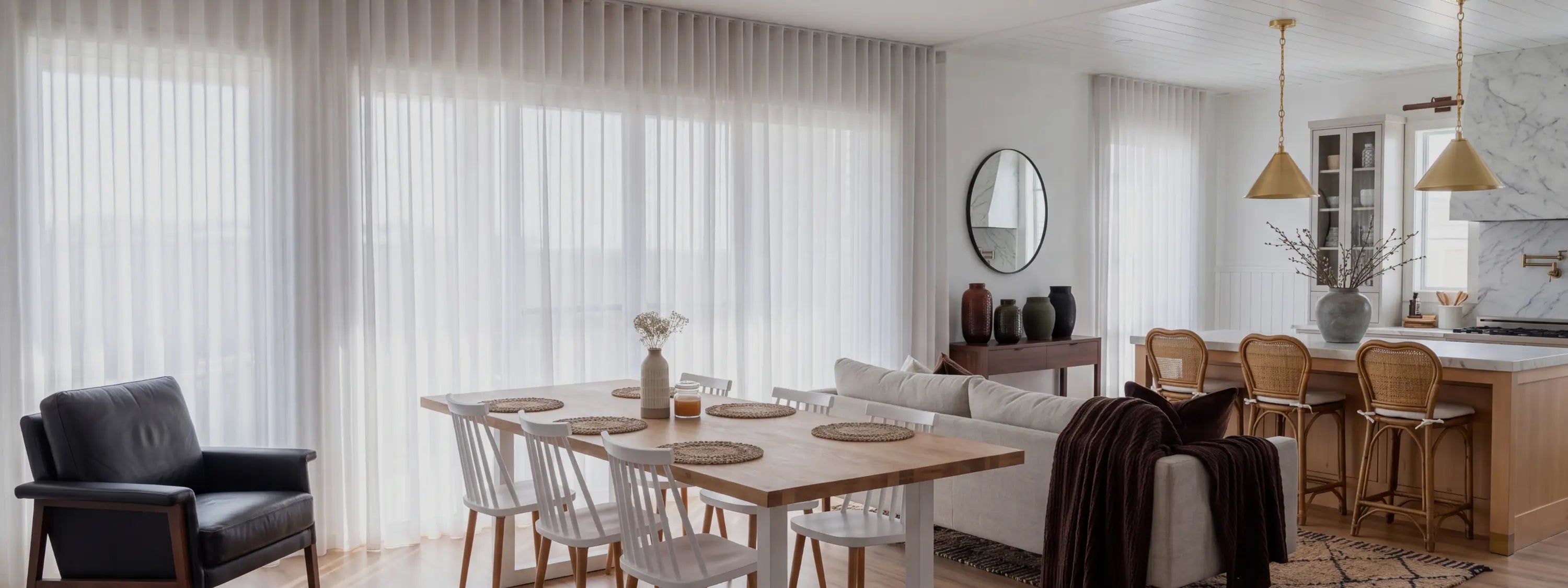 A bright, modern dining room and kitchen interior with tall white vertical blinds covering large windows, a light wood dining table with white chairs, a cream sofa, pendant lights, and a kitchen island in the background.