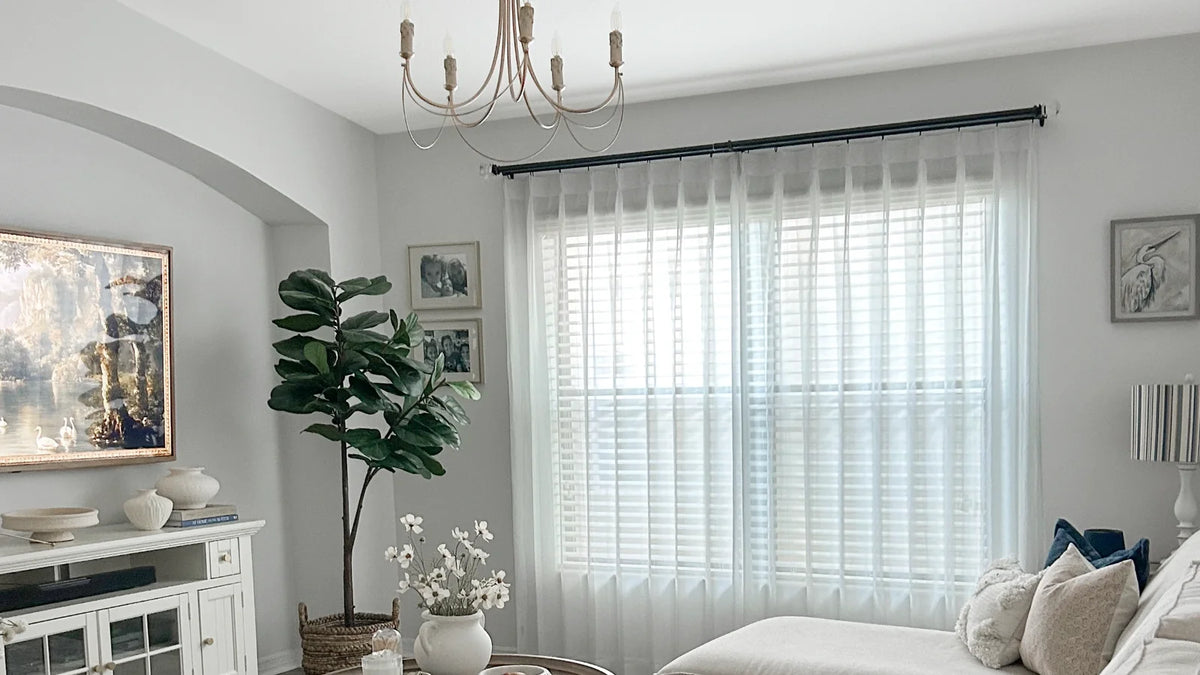 Sheer white curtains on a black rod hung above and outside white horizontal blinds in a living room
