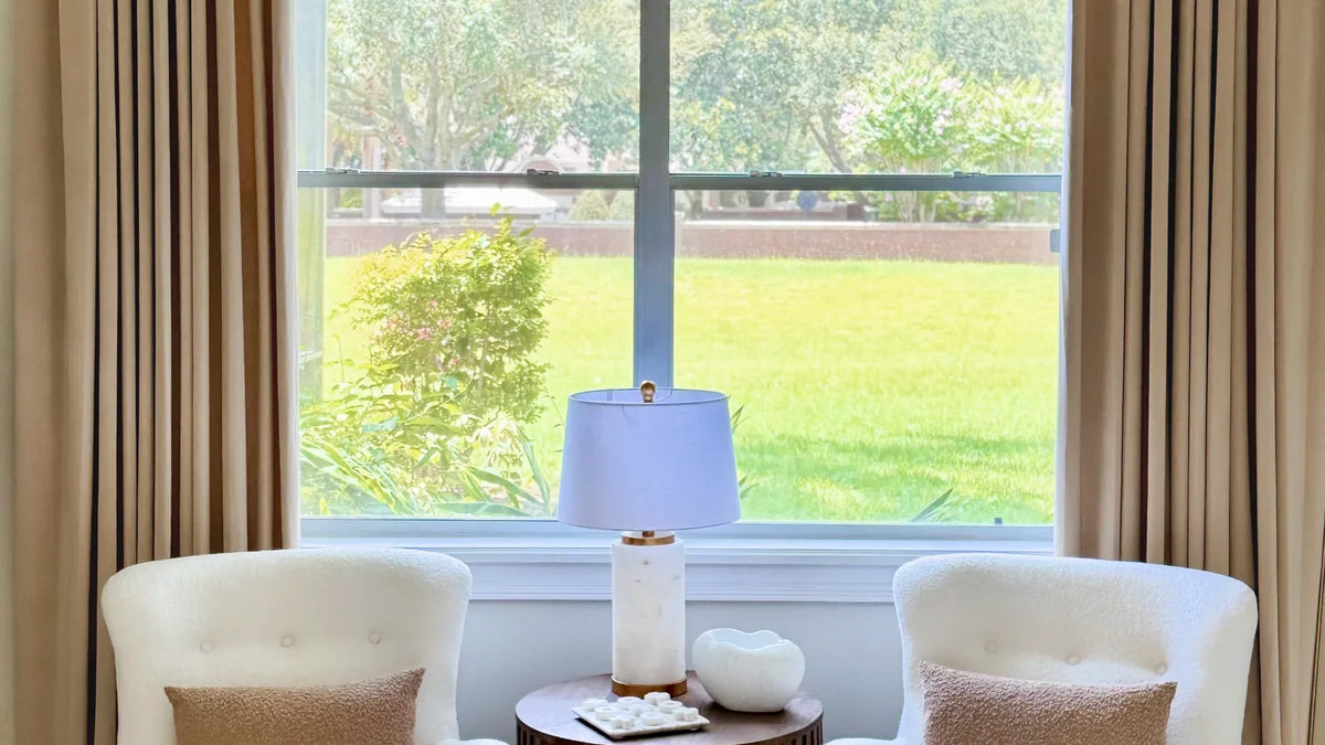 Warm beige velvet curtains framing a sunny window behind two white boucle armchairs and a marble table lamp