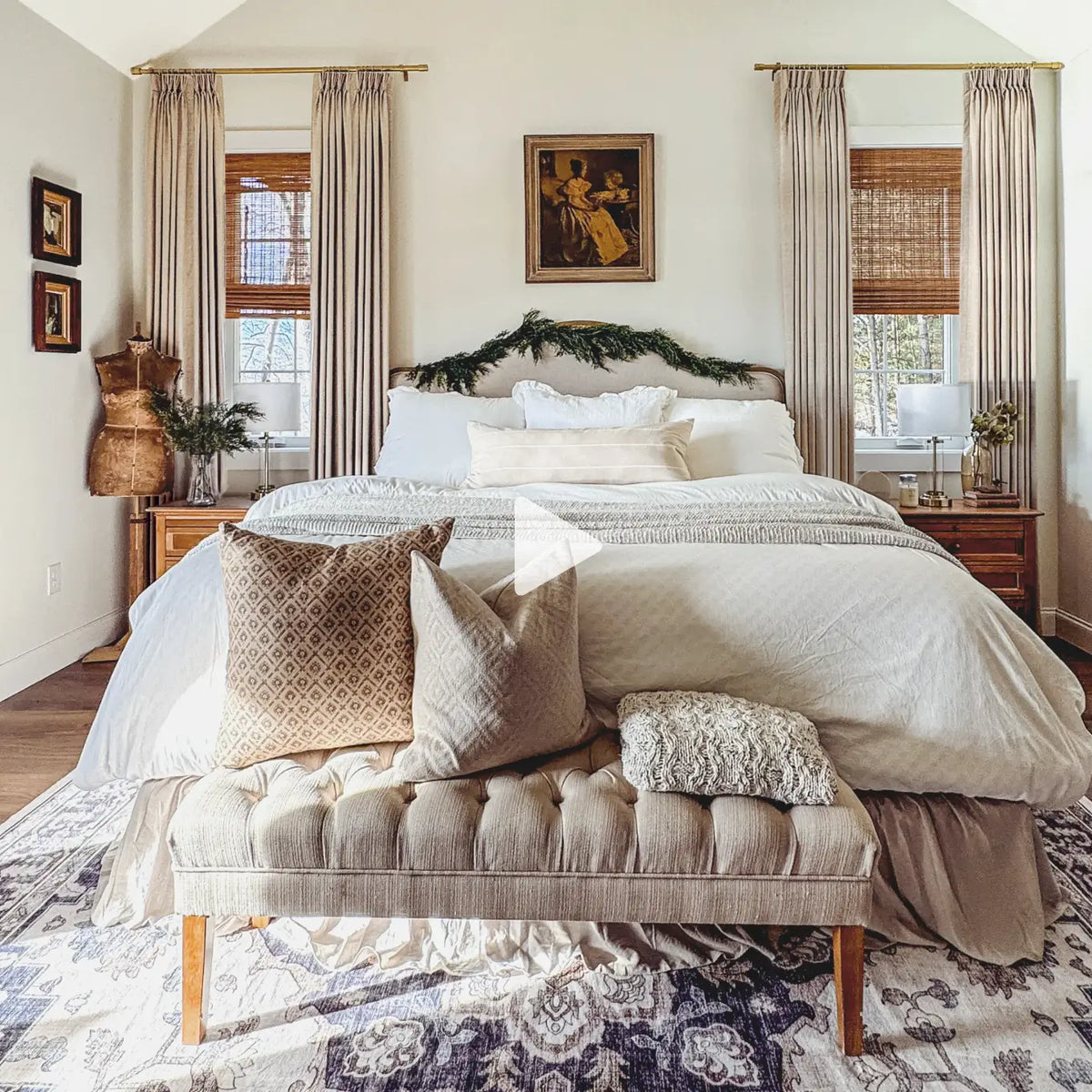 Translation missing: en.A styled bedroom interior with a bed dressed in cream bedding, patterned beige pillows, a tufted bench at the foot of the bed, two tall window treatments with gold rods, and warm wood accents. A large semi-transparent play icon is overlaid in the center of the image