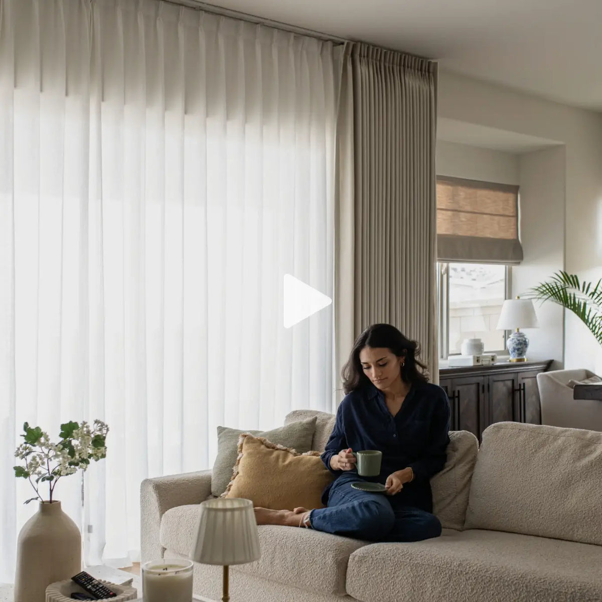 Translation missing: en.A woman sits on a cream bouclé sofa in a bright living room, holding a mug. The room features large white sheer curtains, neutral-toned walls, and tasteful home decor
