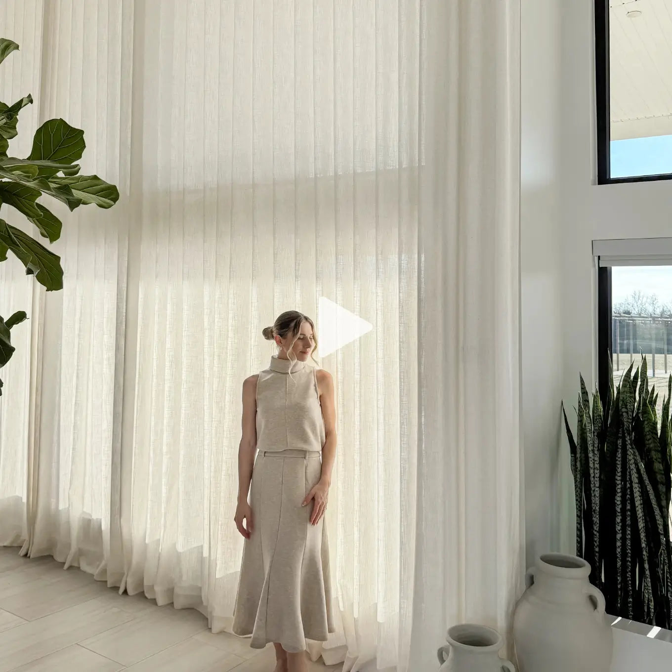 Translation missing: en.A woman stands in a bright room wearing a sleeveless beige high-neck top and matching skirt set. She is positioned in front of large white sheer curtains, with indoor plants and white ceramic vases visible around her