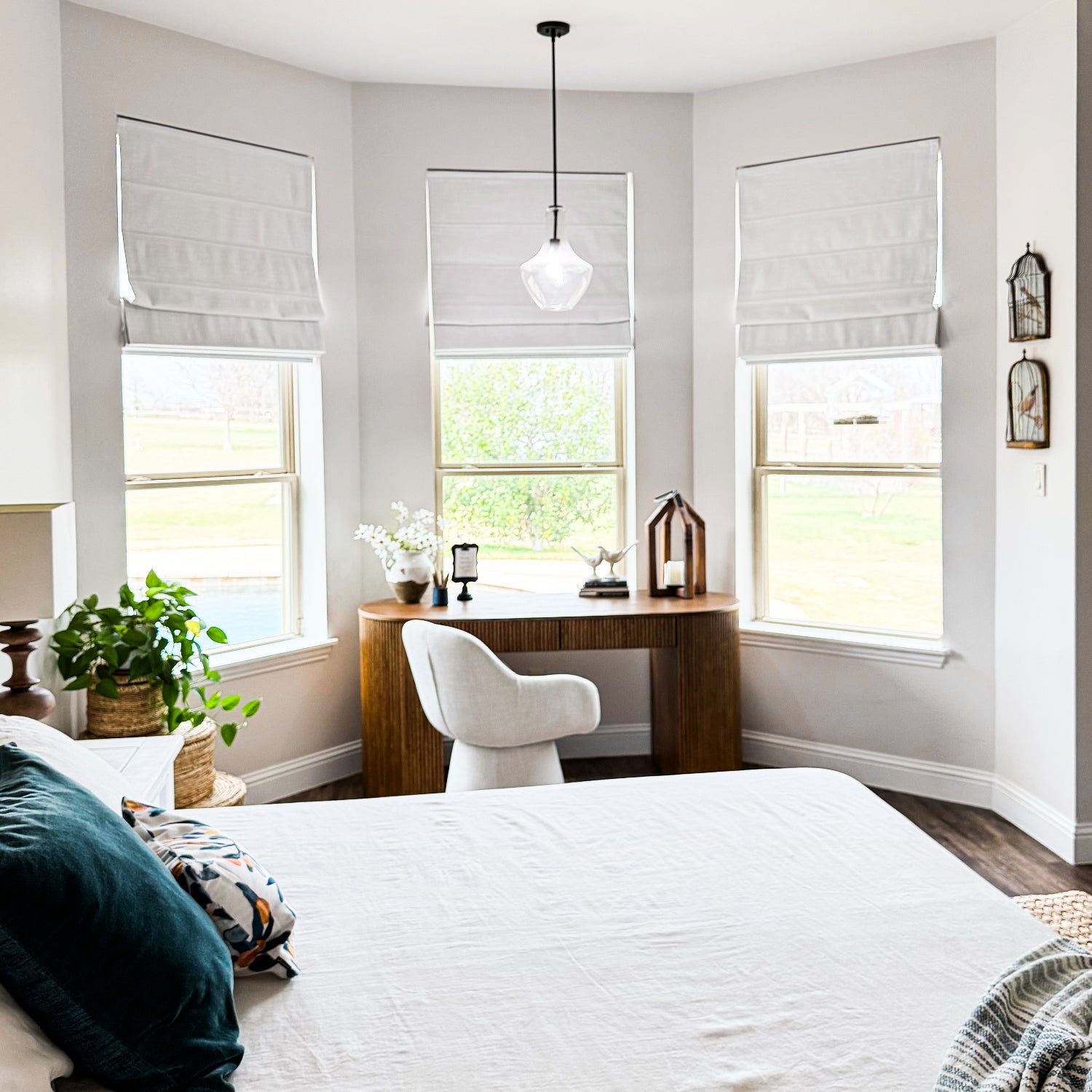 A bright, modern bedroom corner with a bed in the foreground, a wooden desk and white chair by three windows with white roller shades, and a hanging pendant light.
