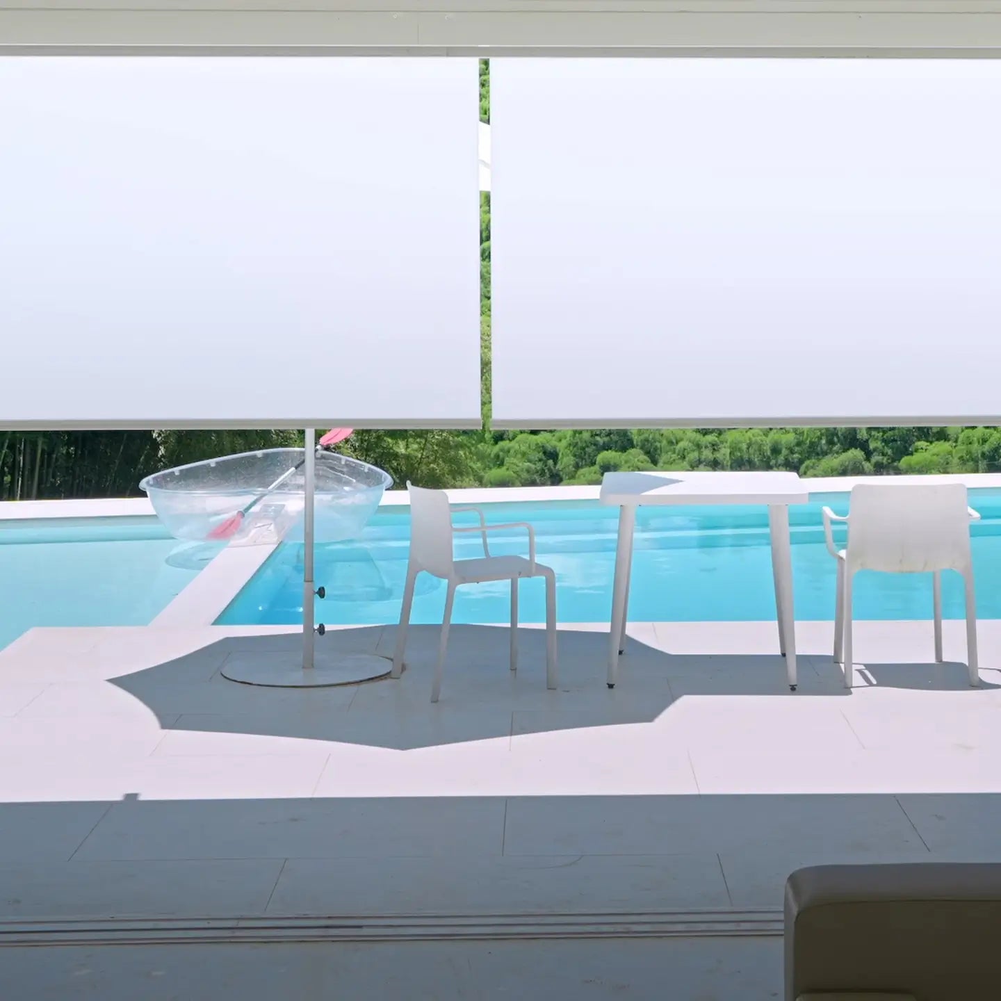 Translation missing: en.Modern outdoor patio furniture set including a white dining table and two matching chairs, alongside a clear round pool float, positioned next to a bright blue swimming pool with a white wall and green trees in the background