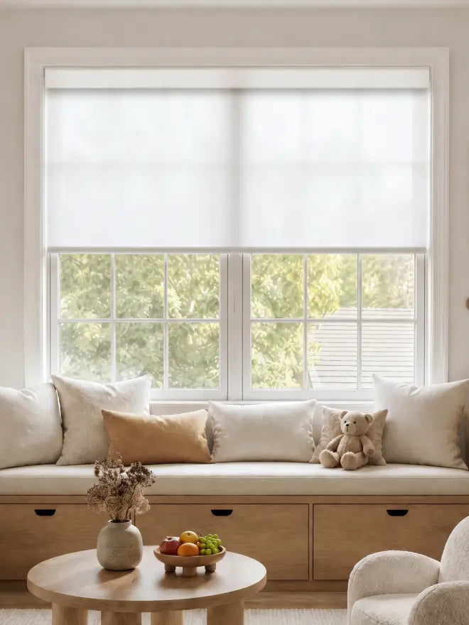A bright, modern room featuring a large window with a white roller blind, a built-in wooden window seat with cream cushions, and a small round wooden coffee table in the foreground.