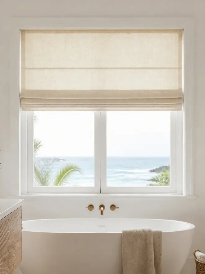 A beige fabric Roman shade drawn over a white window above a white freestanding bathtub with brass fixtures, set in a bright bathroom with an ocean view outside.